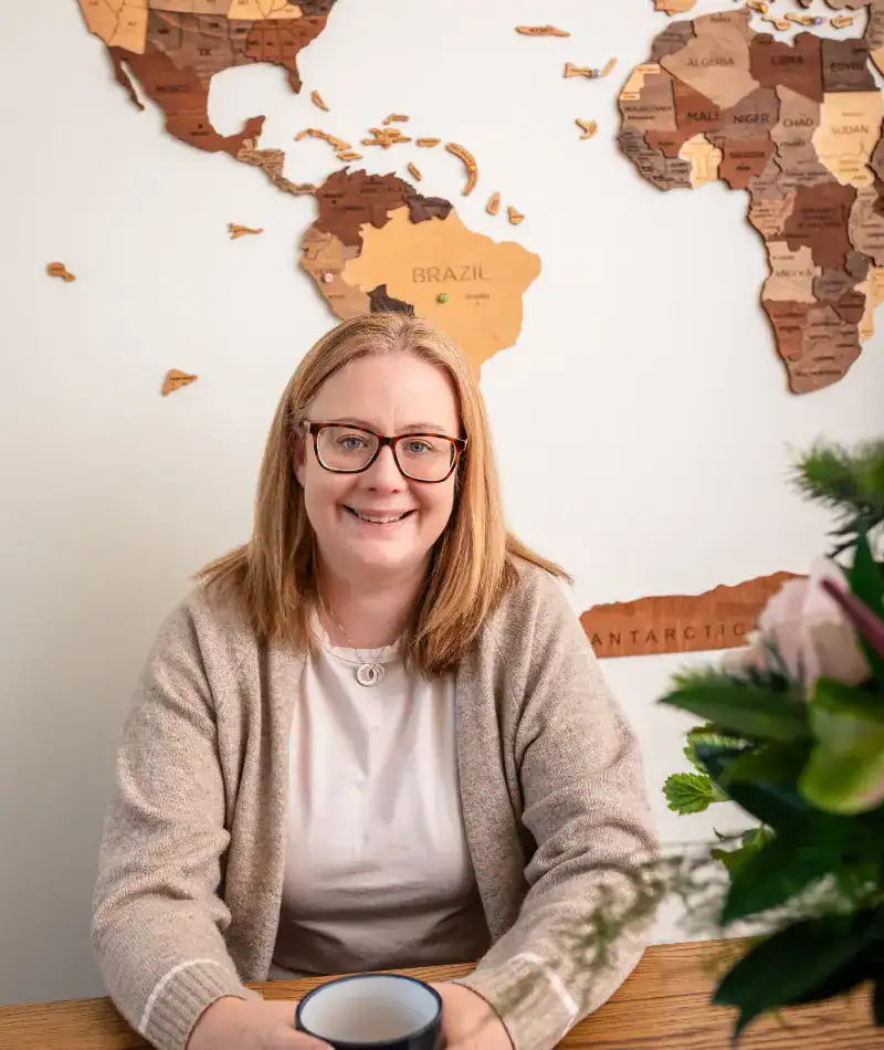 Louise Cook, Galway web designer, smiling at a desk with a coffee and a wooden world map on the wall behind her