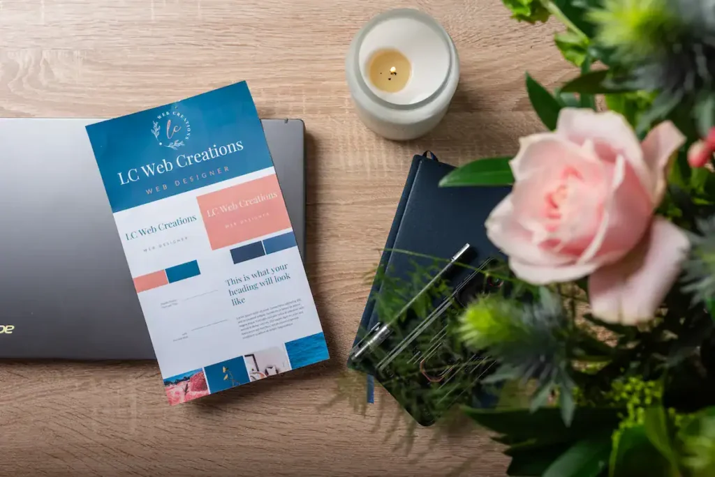 LC Web Creations branded mood board and website design mockup displayed on a desk alongside a laptop, notebook, candle, and fresh flowers