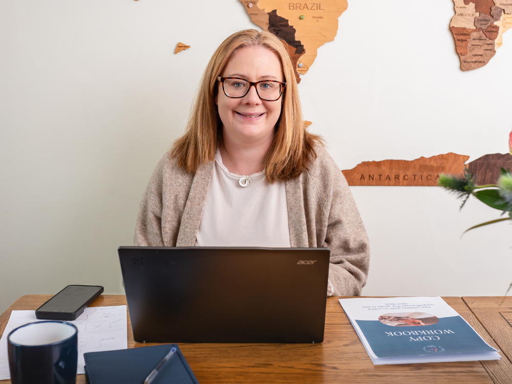 Louise Cook, Galway web designer, smiling at her desk with a laptop, fresh flowers, and a wooden world map on the wall behind her