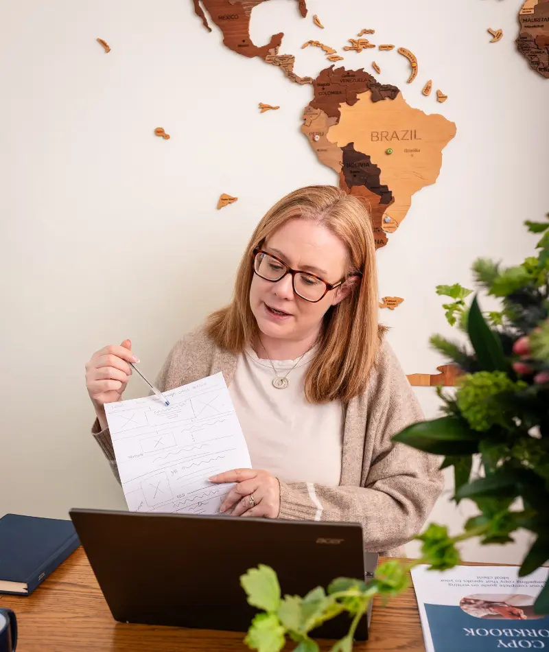 Louise, Web designer in Galway, sitting at a table explaining the website audit to someone on the open laptop. World map shown in the background