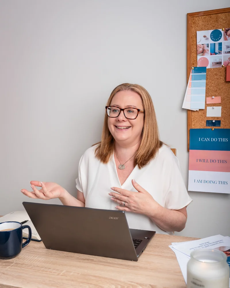 Louise, web designer in Galway, sitting at her desk with laptop open giving feedback of a website audit. Her hands are up and she's smiling. Noticeboard with branding on the wall